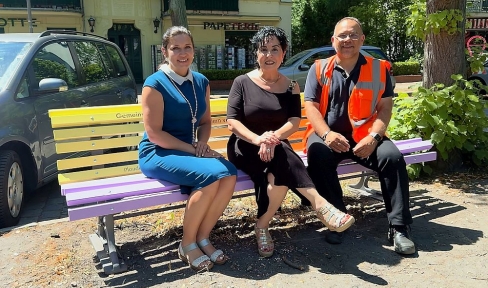 Zwei Frauen im kurzen Kleid und ein Mann mit Warnweste sitzen auf einer gelb-lila lackierten Bank.